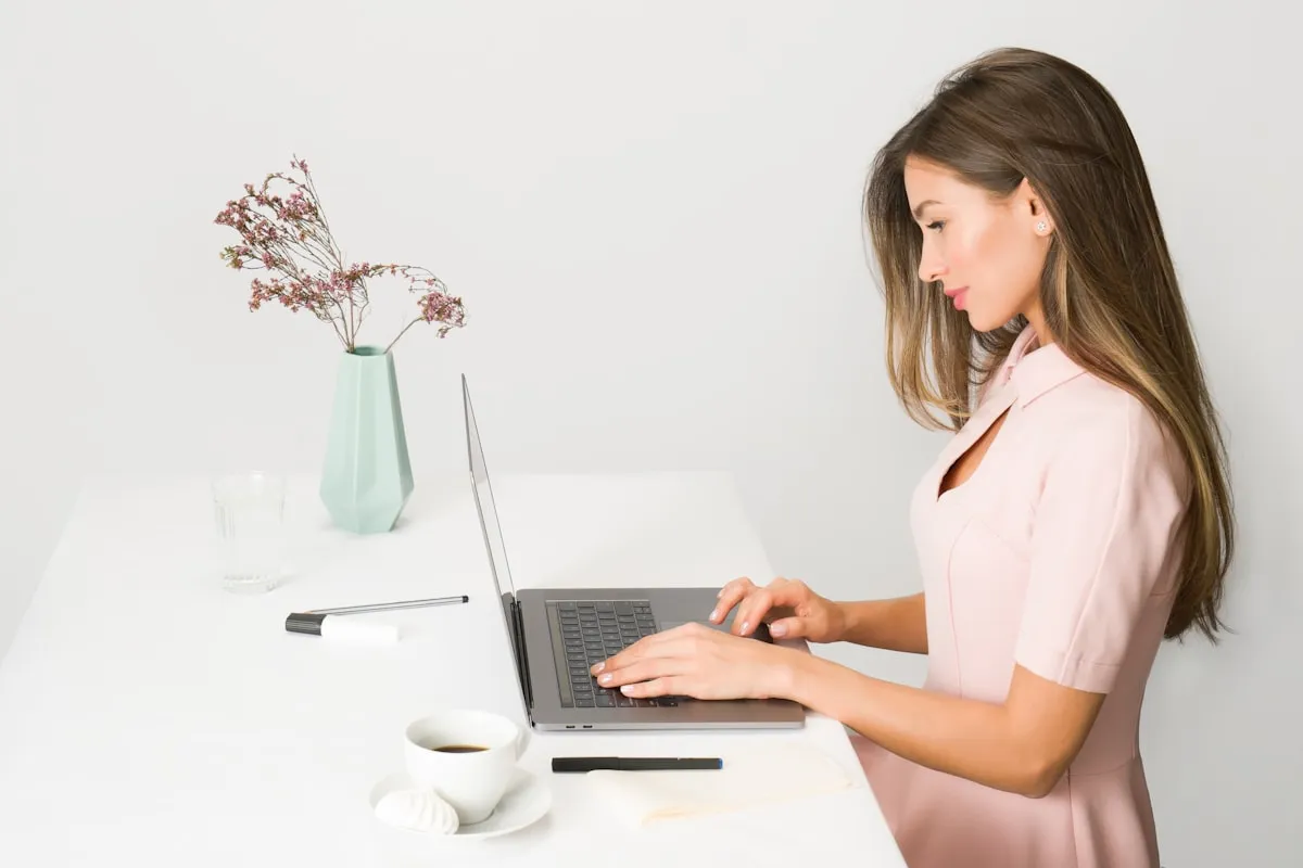 A maker working at a laptop on a bright white desk, with a notebook, pen, and a cup of coffee beside the keyboard.