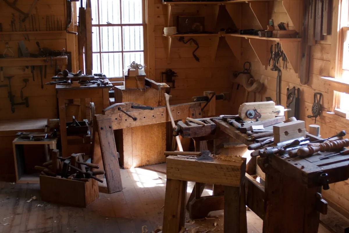 An empty timber-framed woodworking shop with hand tools racked on the walls, planes and chisels laid out across two workbenches, and sunlight coming through the window