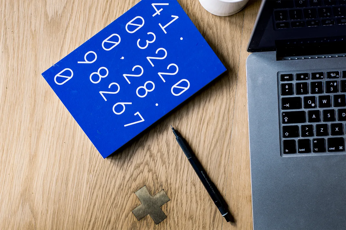 An overhead flat-lay of a wooden desk: a blue notebook covered in large printed numbers sits beside the edge of a laptop and a black pen