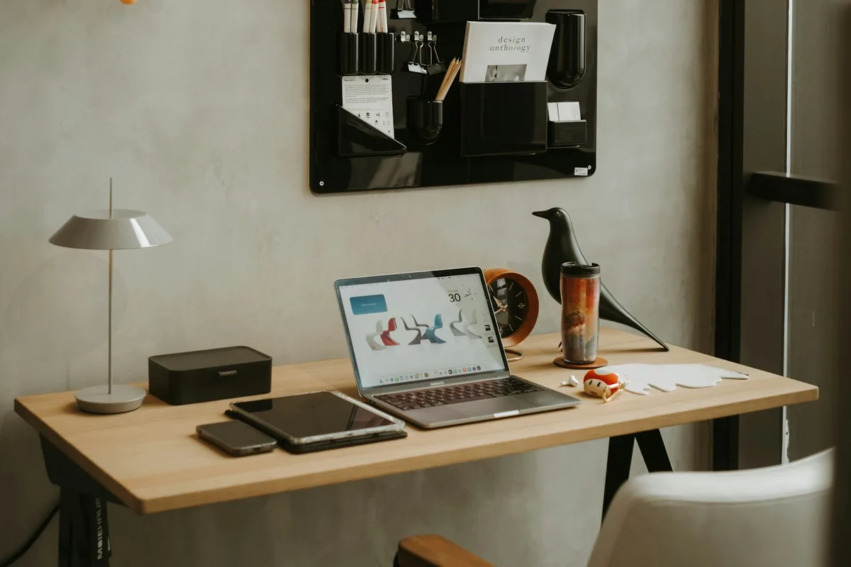 A small-business workspace with a laptop open on a wooden desk, a phone and tablet alongside it, a wall organizer holding design tools and a notebook above the desk, and a desk lamp and tumbler completing the setup