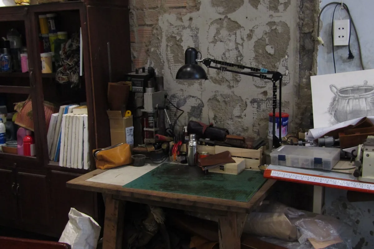 An empty leather workshop bench with a green cutting mat, an anglepoise lamp, hand tools and offcuts, with a pencil drawing of a bag propped against the wall behind it