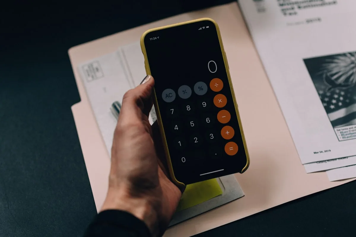 A hand holding a smartphone calculator over a manila folder of printed paperwork on a dark desk — working out a price