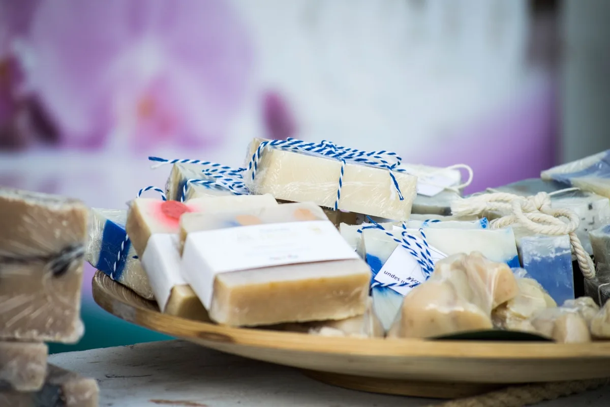 A wooden tray heaped with handmade soap bars wrapped in cellophane, each tied with blue-and-white baker's twine and carrying a small paper band label or hang tag, with a printed white wrap label facing the camera