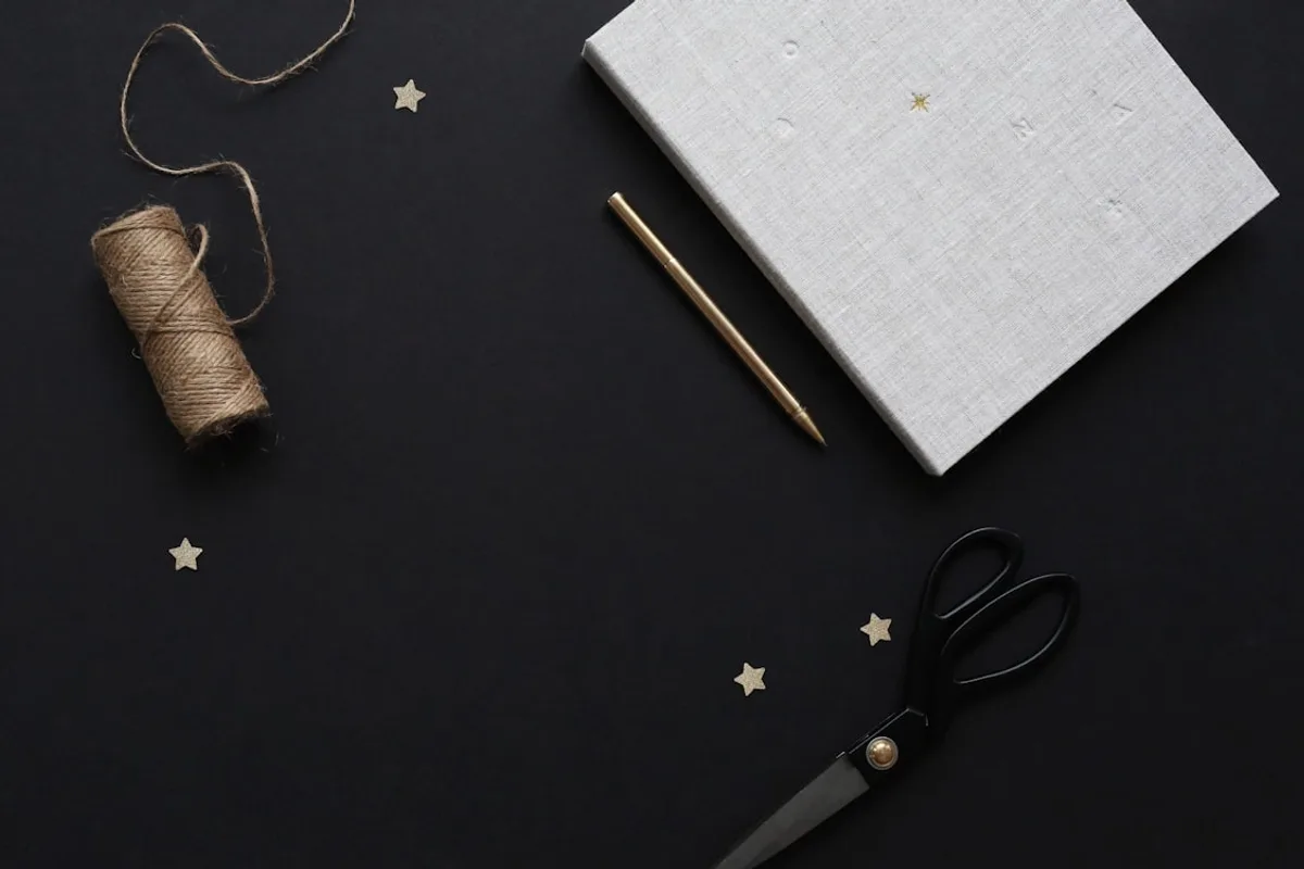 A handmade seller's planning flat-lay on a dark surface — a linen notebook, a gold pen, a spool of twine, tailor's scissors, and scattered gold star confetti