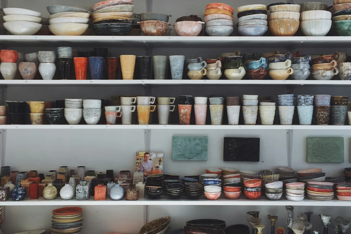 Rows of white shop shelving closely stacked with handmade glazed ceramics — bowls, tumblers, mugs and small vases in mixed colors and finishes