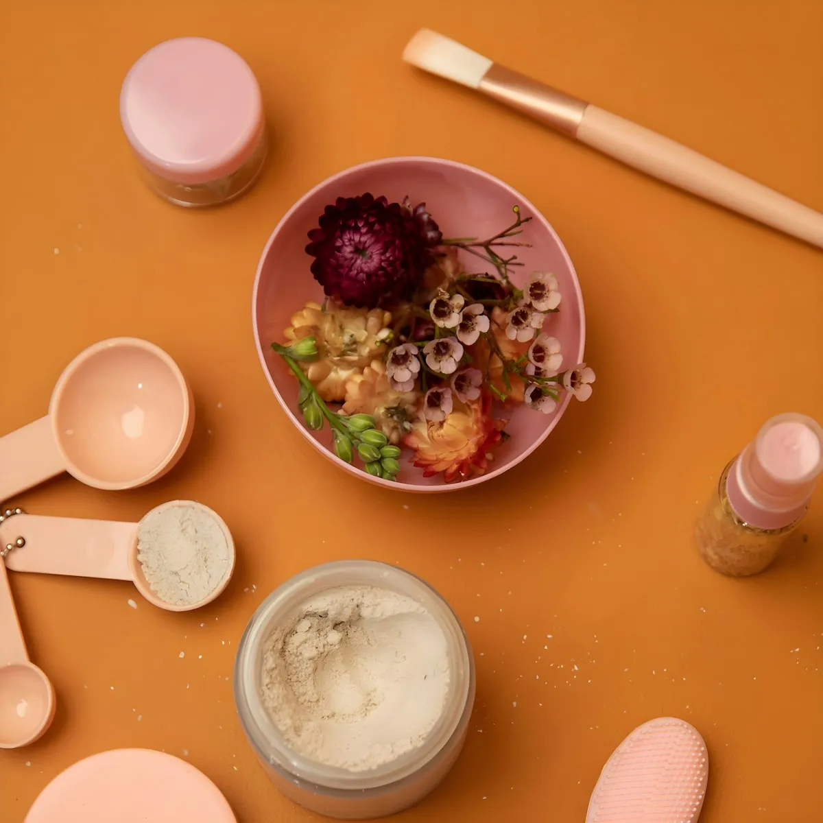 Flat lay of skincare making ingredients and tools including measuring spoons, powder jars, a dropper bottle, mixing brush, and dried flowers on an orange surface