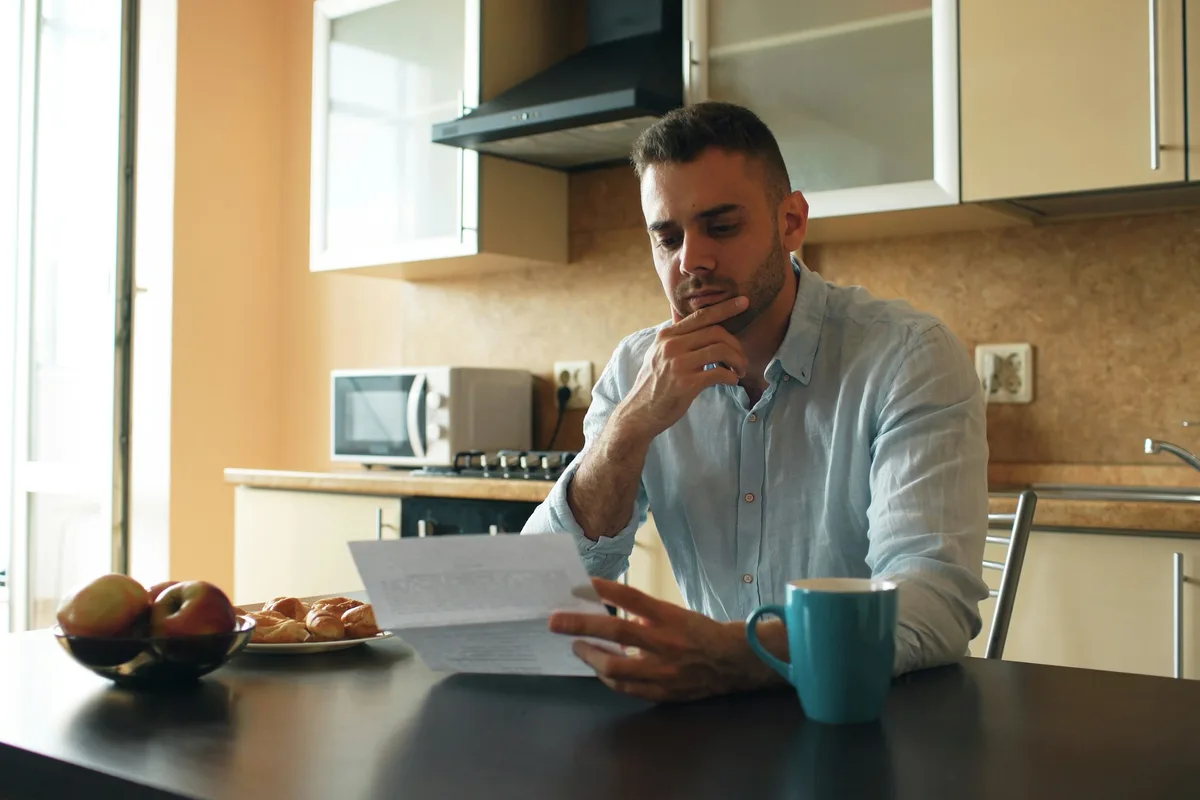 A person sitting alone at a kitchen table in morning light, chin in hand, reading a single printed document beside a mug of coffee and a bowl of fruit