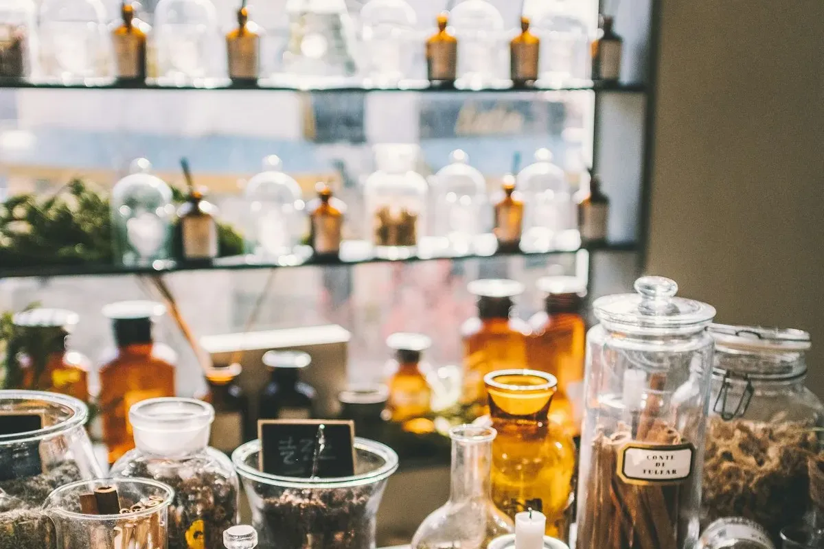 A sunlit apothecary workspace with amber tincture bottles on glass shelves and dried herbs, cinnamon sticks, and botanical ingredients in bowls and jars on a wooden surface