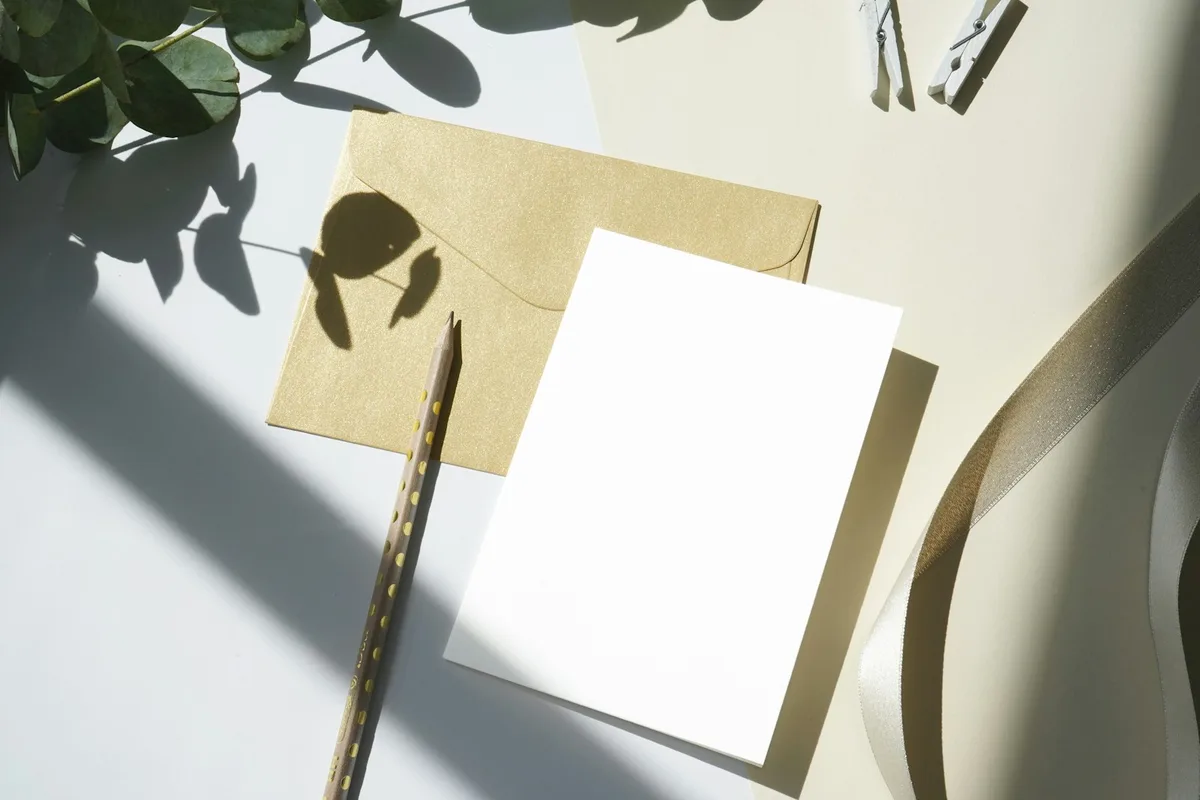 A sunlit flat-lay of a blank cream greeting card and a gold kraft envelope on a pale surface, with a spotted pencil, a coil of satin ribbon, wooden clothespins, and a sprig of eucalyptus casting soft shadows