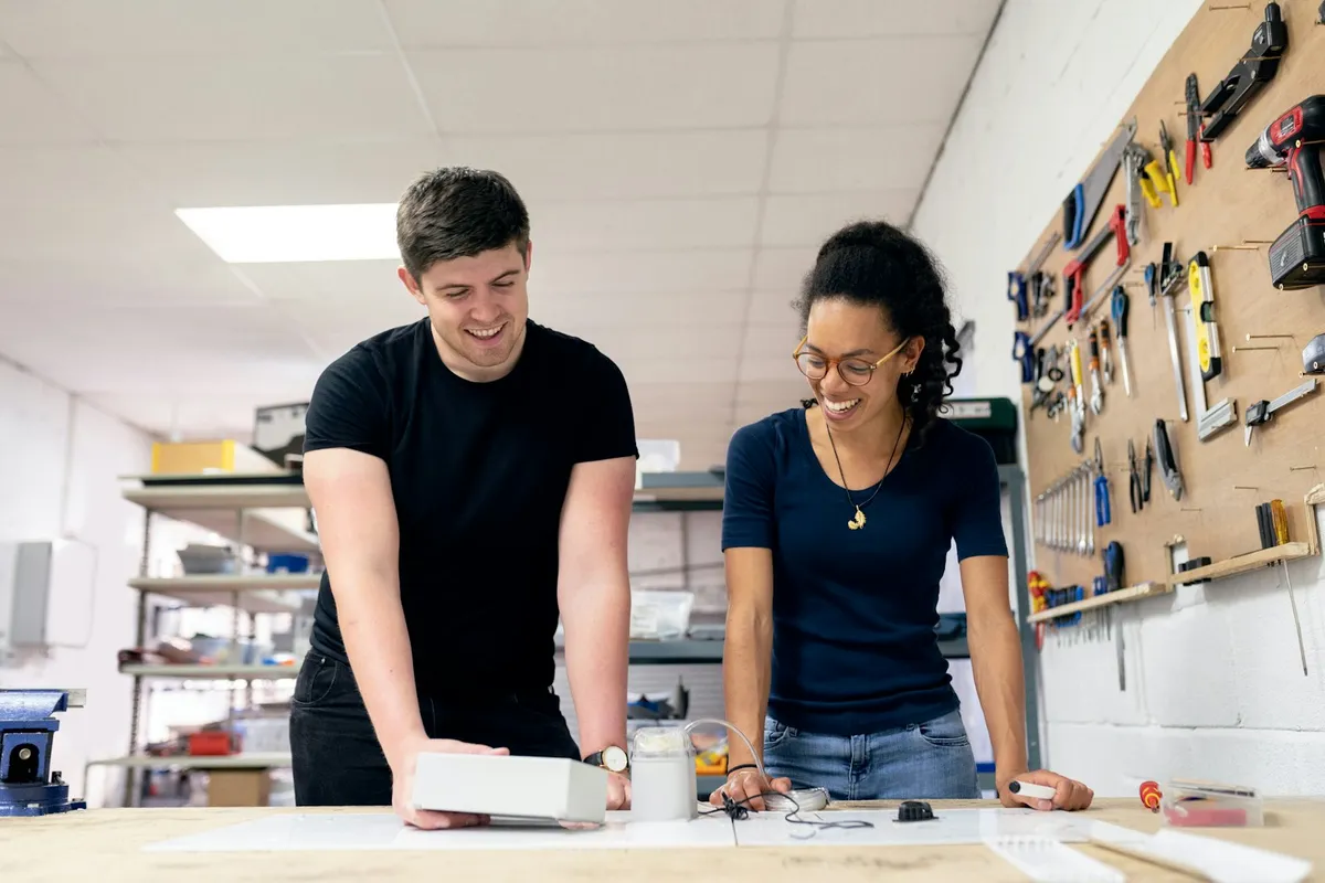 A maker and a helper smiling as they examine a small device together at a workshop bench, with a pegboard wall of organized hand tools and parts shelving in the background