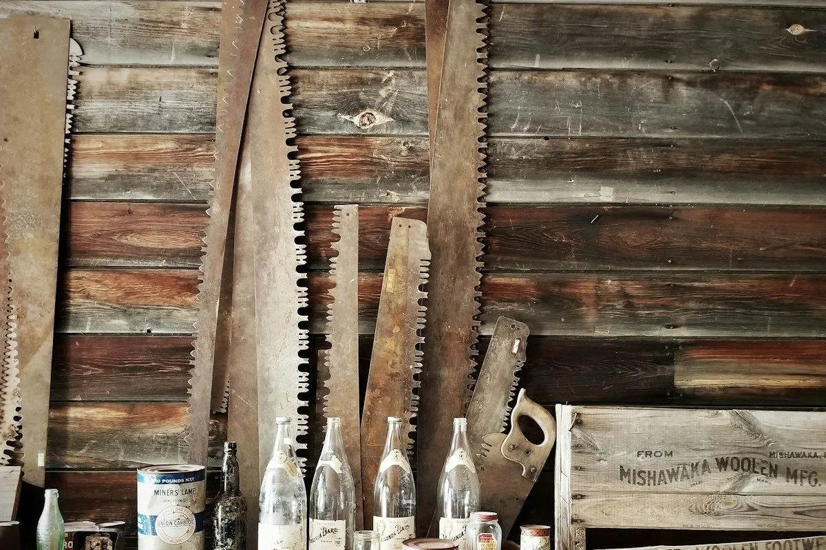 A weathered wooden shop wall hung with antique handsaws, faded glass bottles on a shelf, and a stenciled wooden crate reading "Mishawaka Woolen MFG" — a vintage maker workshop scene