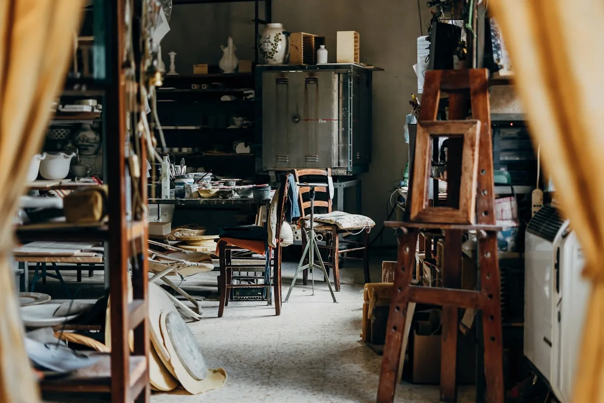 A working artisan pottery studio seen through a doorway — crowded shelves of work in progress, a kiln, a wooden ladder, and finished pieces filling a busy maker workspace