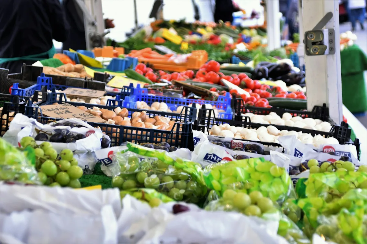 A long farmers market vendor table piled with crates of grapes, mushrooms, tomatoes, peppers, and seasonal produce, with the vendor visible in the background
