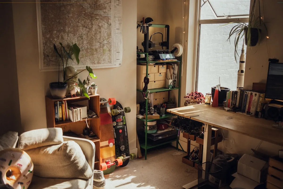 A sunlit room that is half living space and half workshop — an armchair and bookcase on one side, a workbench, parts shelving and a small 3D printer along the window wall