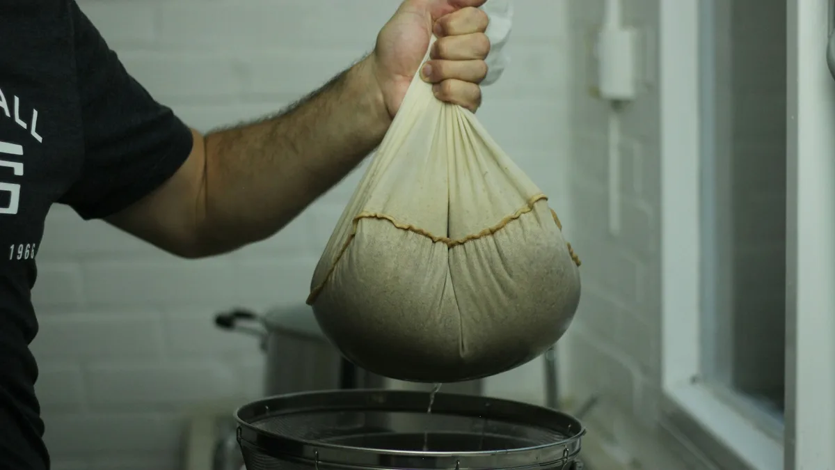 A food producer straining liquid through a muslin bag into a stainless steel stockpot inside a clean white-tiled production kitchen