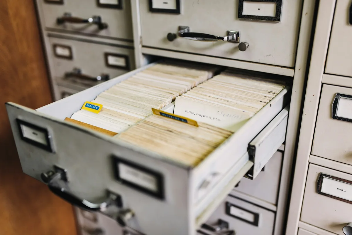 An open drawer in a gray metal filing cabinet, packed with rows of aged paper index cards behind colored divider tabs