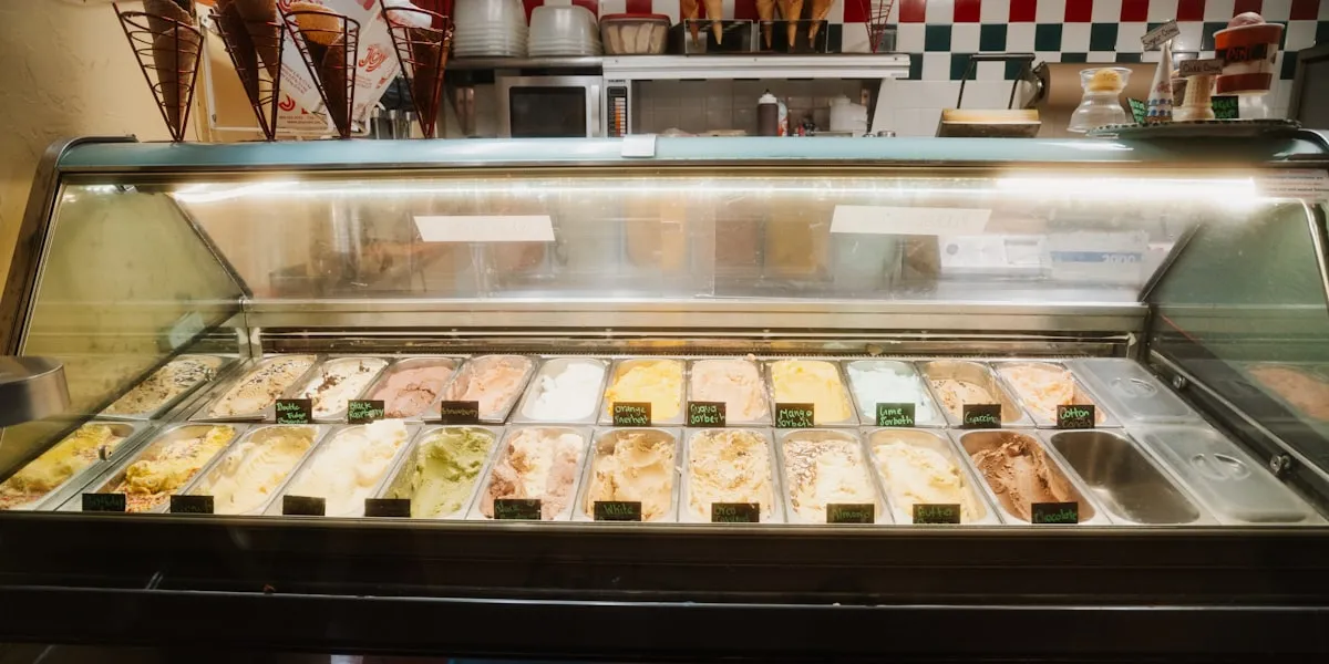 Gelato display case filled with labeled flavors in metal pans, waffle cones overhead, in a gelateria with checkered tile walls