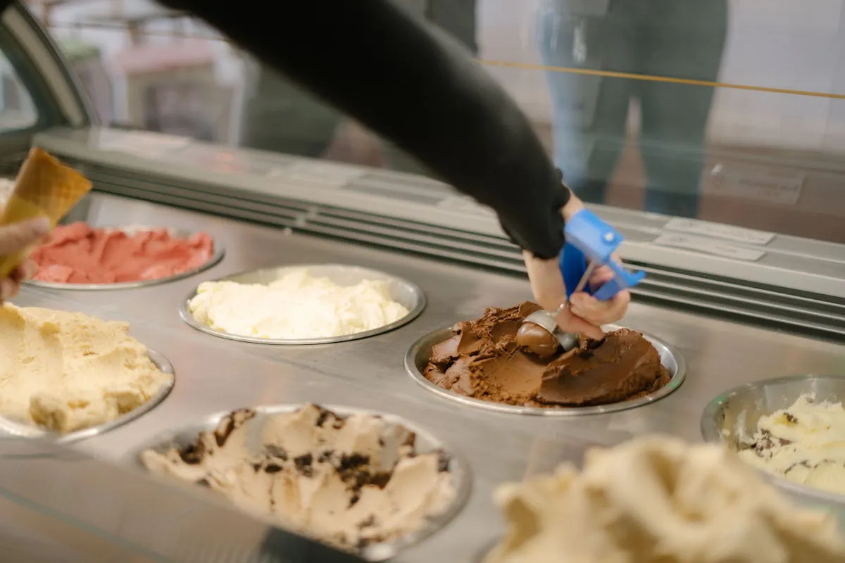 Person scooping chocolate gelato from a colorful display case of assorted artisan gelato flavors in a bright shop