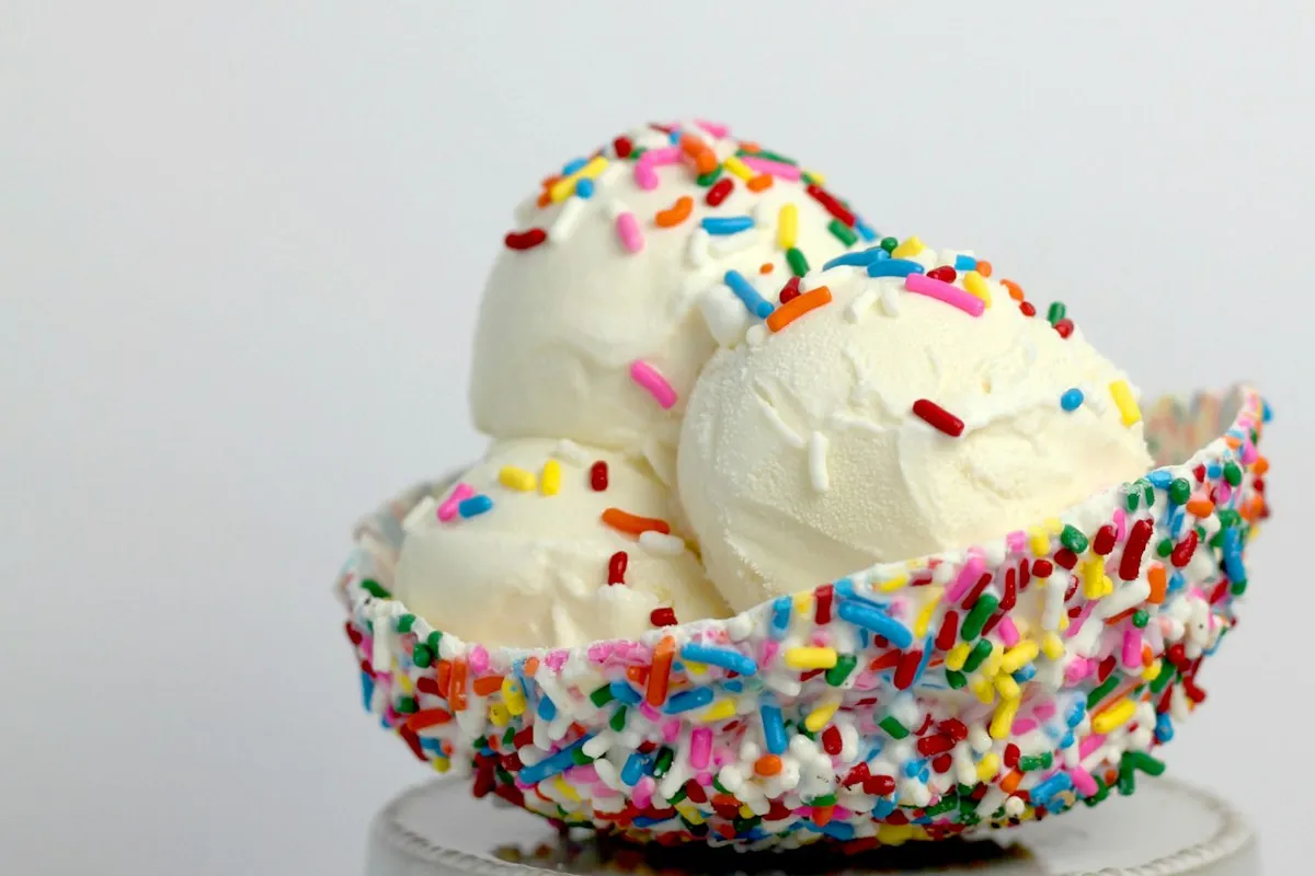 Scoops of vanilla ice cream topped with rainbow sprinkles in a sprinkle-rimmed waffle bowl on a small white stand