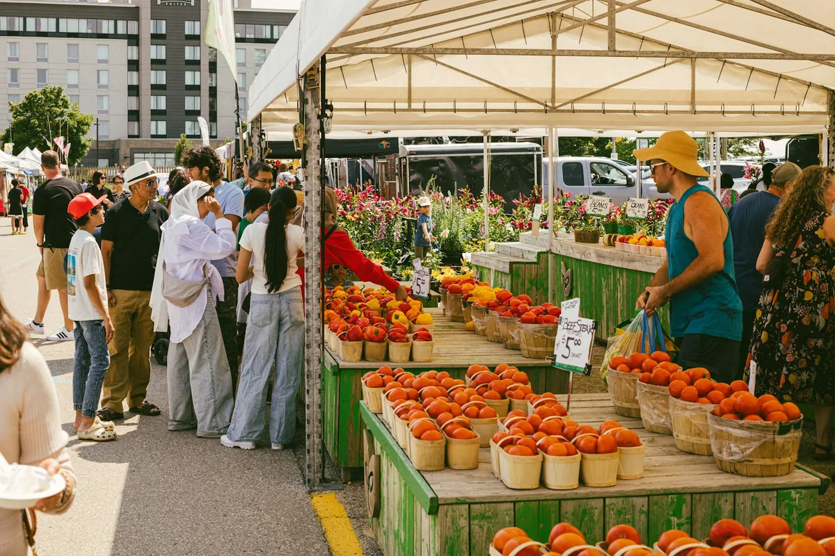 Customers browsing baskets of fresh peaches and tomatoes at a green wooden produce stall at an outdoor farmers market