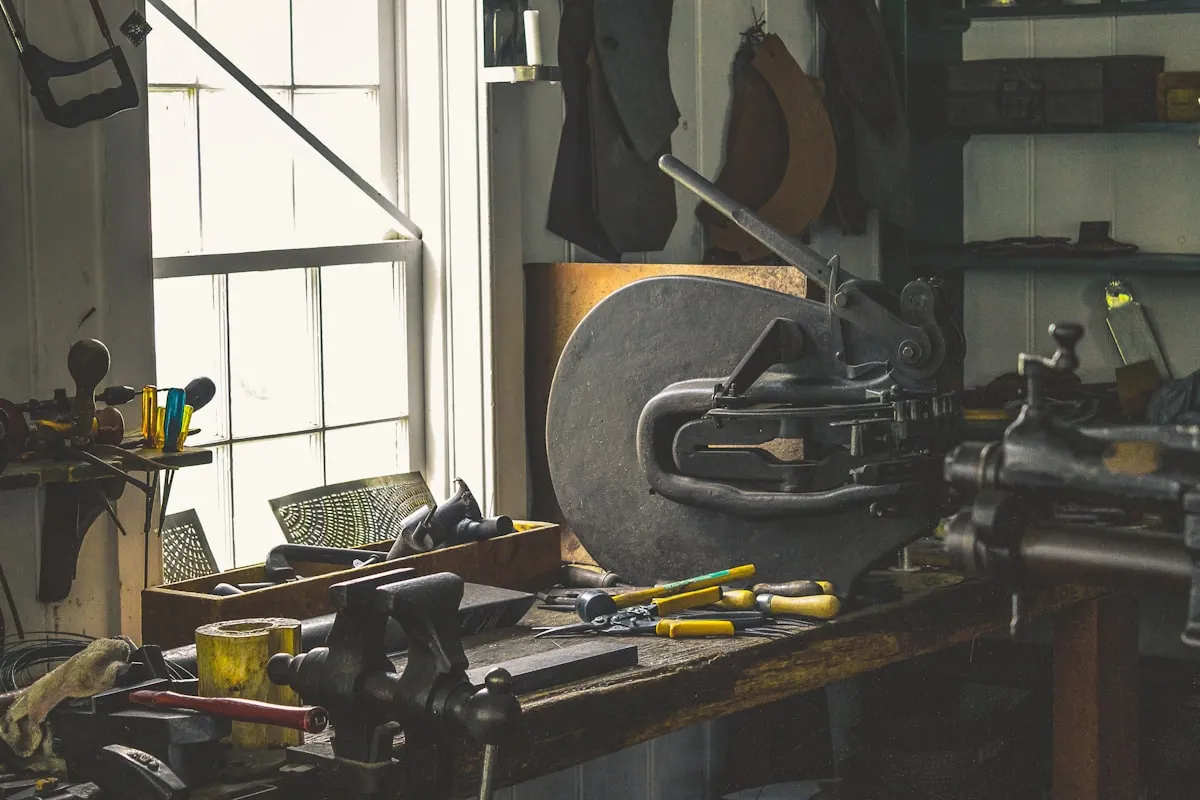A sunlit workbench in a small workshop holding a bench vise, screwdrivers, pliers, and a cast-iron bench machine, with hand tools hanging beside a paned window