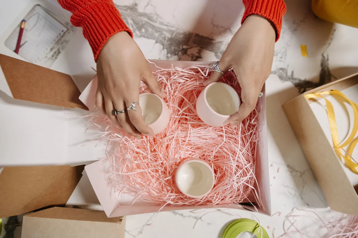 Overhead view of hands in a red sweater setting three small pink ceramic cups into a gift box lined with pink shredded paper, with kraft boxes and ribbon on the table around it