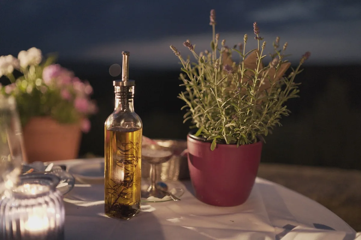 A tall glass bottle of rosemary-infused olive oil on a linen-covered table at dusk, beside a potted lavender plant and a lit candle