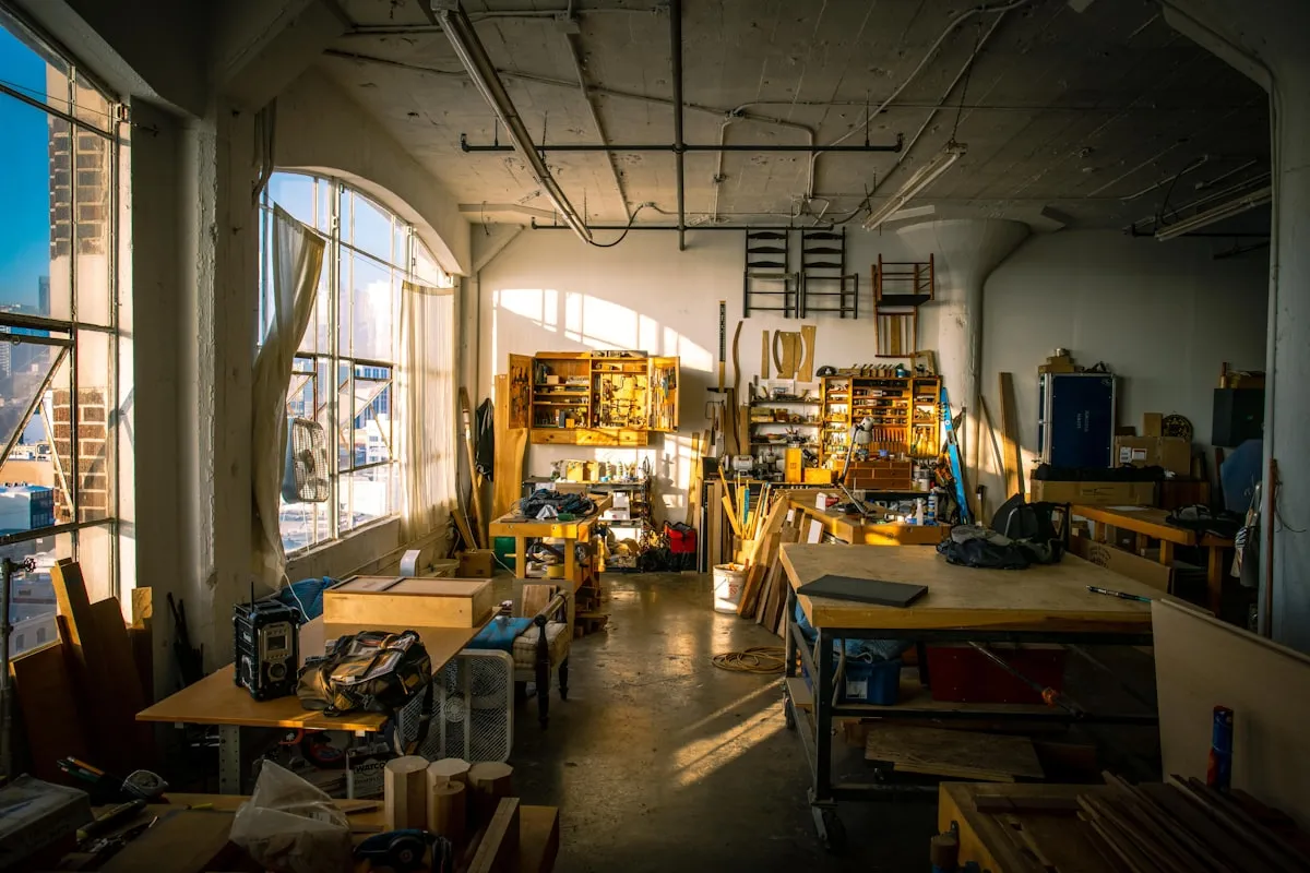 A long sunlit maker's workshop interior with multiple wooden workbenches receding into the distance, a wall-mounted tool cabinet, hanging chairs, scattered offcuts of lumber, and bright morning light streaming through tall arched windows on the left