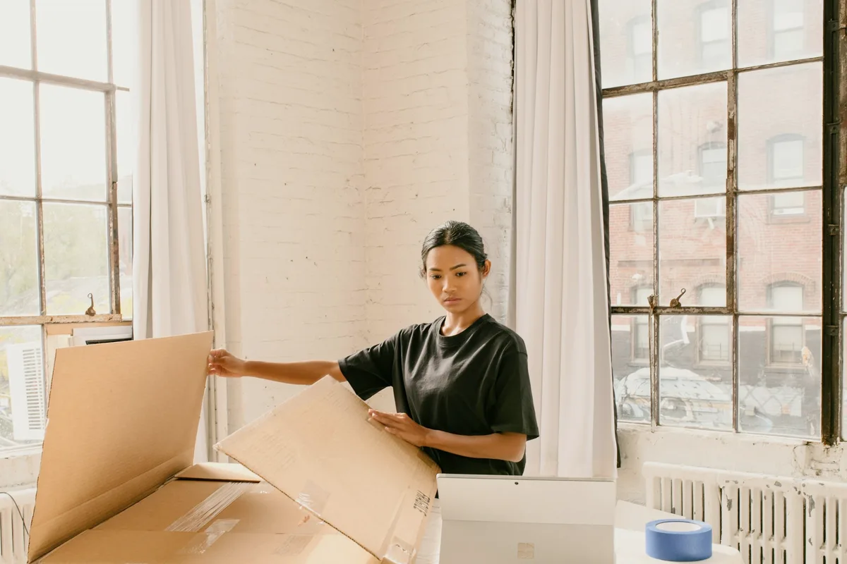 A woman in a black T-shirt folds open a large cardboard shipping box beside a laptop in a bright studio with tall factory windows and white brick walls