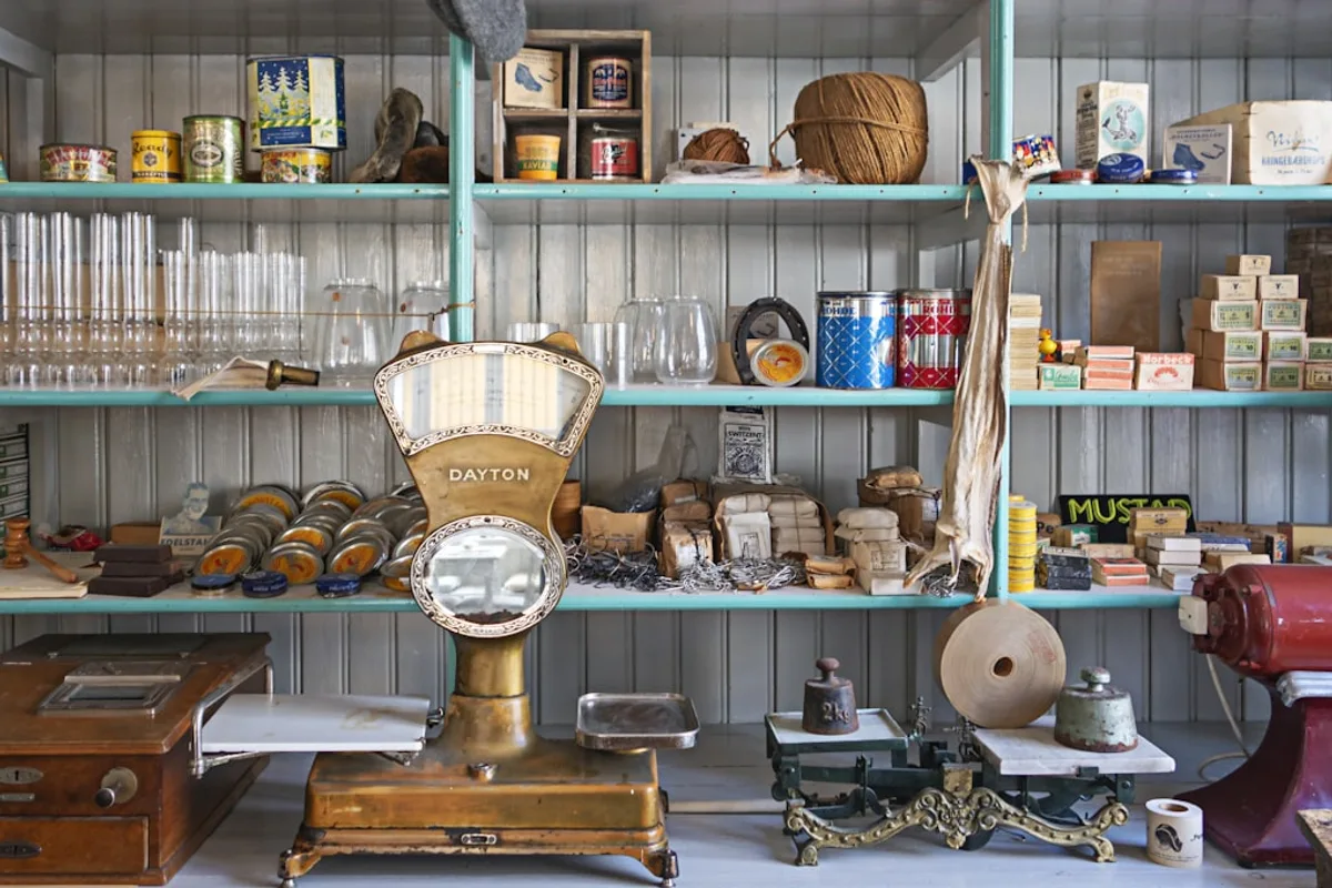 Shelves of a vintage general store stacked with countable tins, boxes and glassware, with two balance scales and a two-kilogram weight on the counter below them