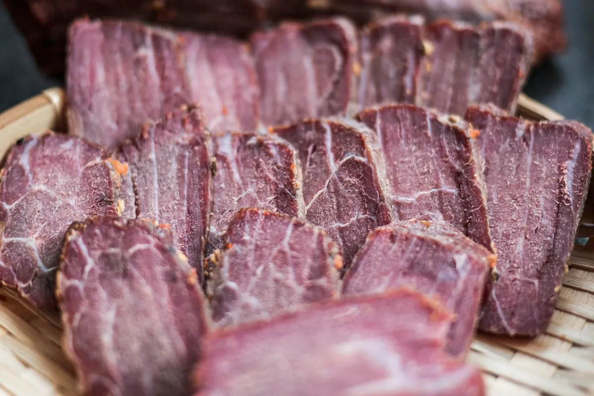 Slices of dark, dried cured beef with deep red marbling arranged in a woven bamboo basket — finished jerky-style dried meat