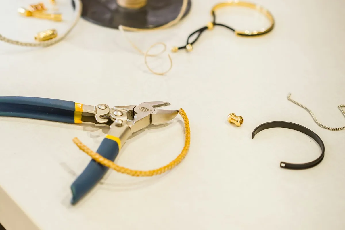 A jeweler's bench with chain-nose pliers, a gold chain, gold and black cuff bracelets, and loose findings arranged on a pale work surface