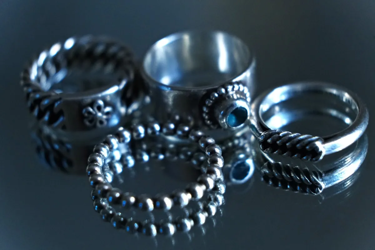 Collection of artisan silver rings with twisted, beaded, and smooth textures and small gemstone settings on a dark reflective surface