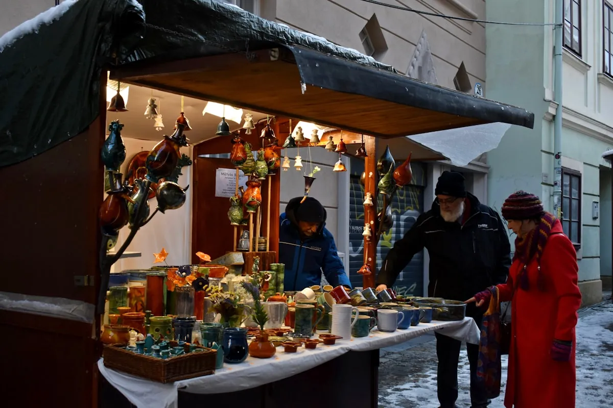A handmade-pottery vendor stands behind a winter market booth filled with brightly glazed mugs, bowls, and hanging ceramic ornaments while two customers in winter coats browse the table