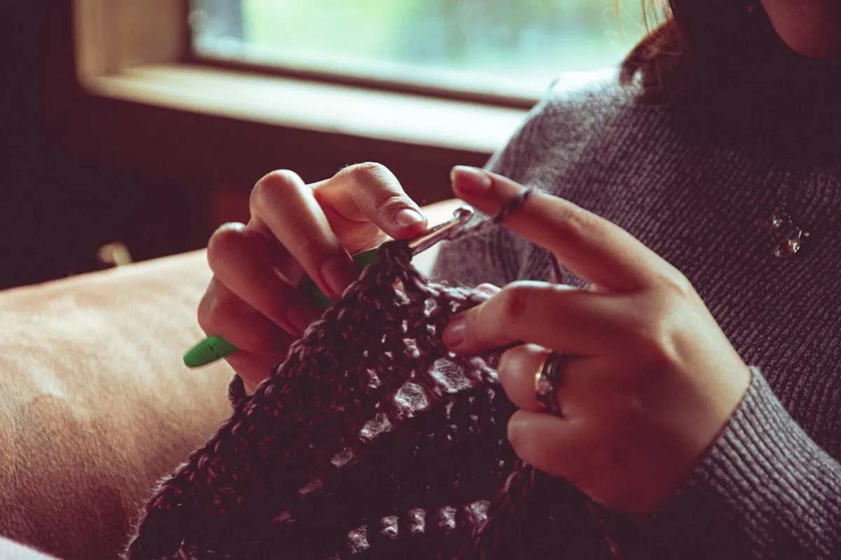 Close-up of hands crocheting a dark textured piece with a green hook in soft natural window light