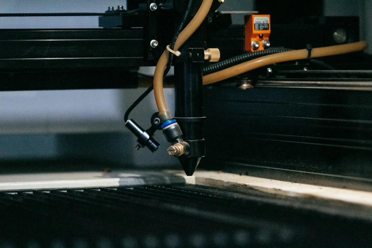 Close-up of a CO2 desktop laser cutter head — air-assist nozzle, brass nozzle tip, flexible exhaust tube, and gantry — poised above the honeycomb bed of a small-workshop machine