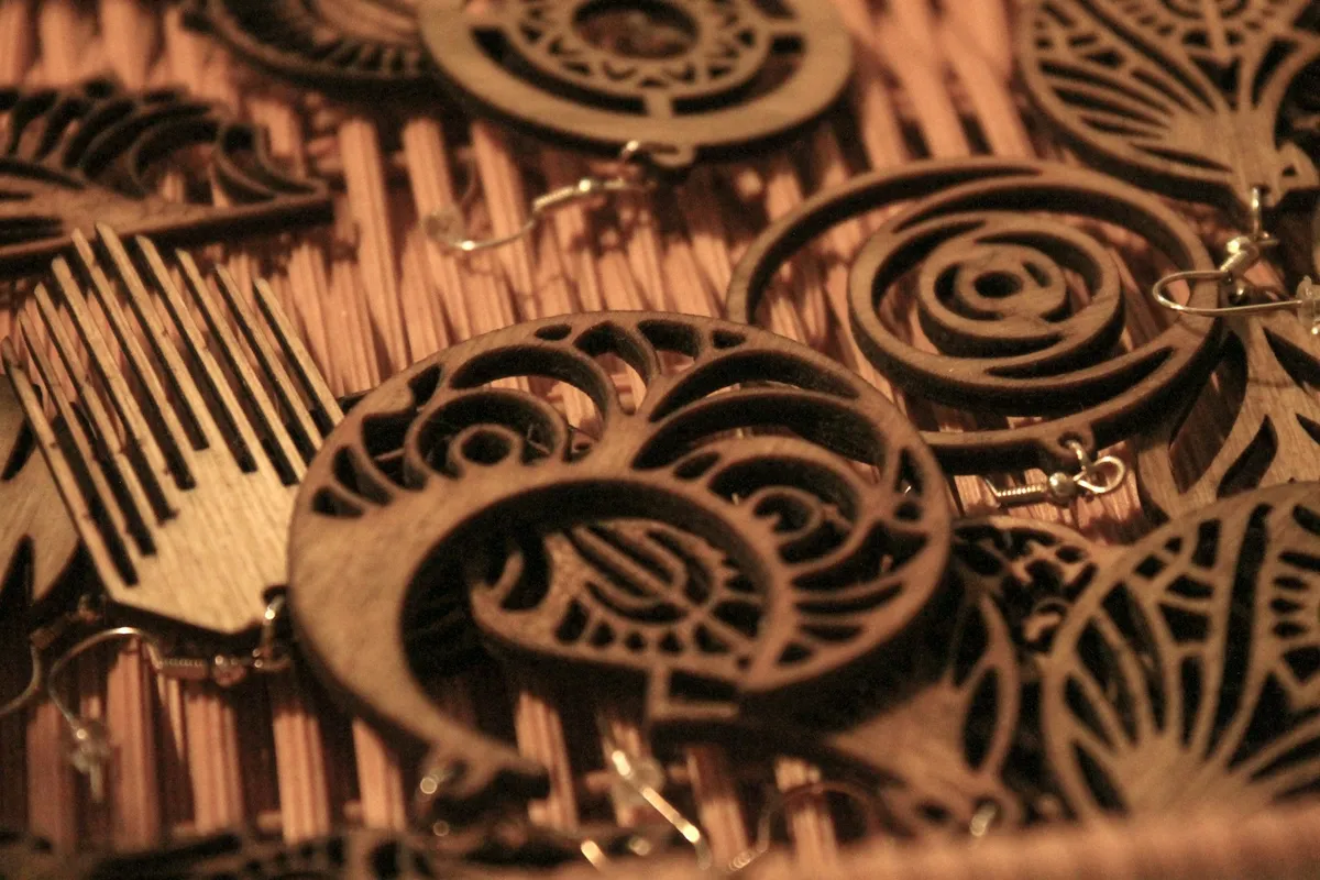 A close-up pile of intricate laser-cut wooden earrings with silver ear wires, their scrollwork and leaf cutouts casting shadows on a woven mat