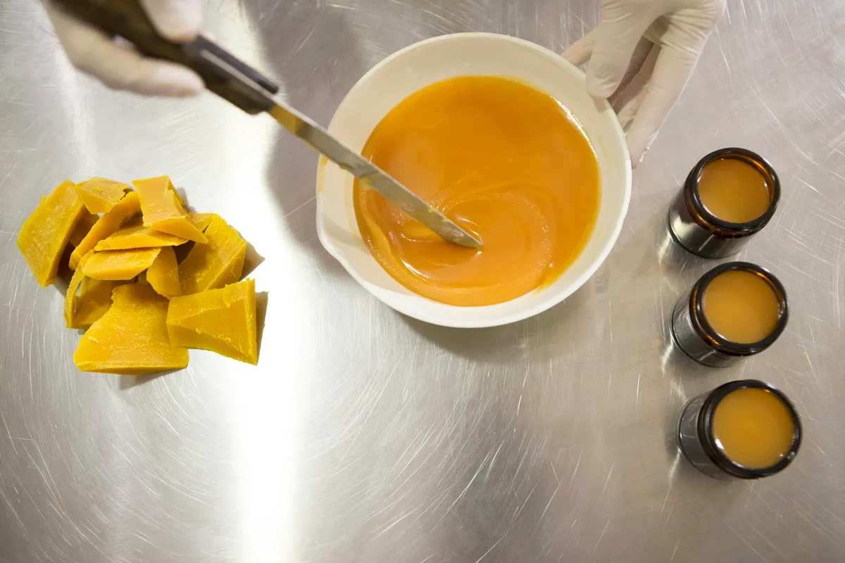 Gloved hands stirring a bowl of melted golden balm with a spatula on a steel workbench, beside a pile of raw yellow beeswax chunks and three small amber jars already filled