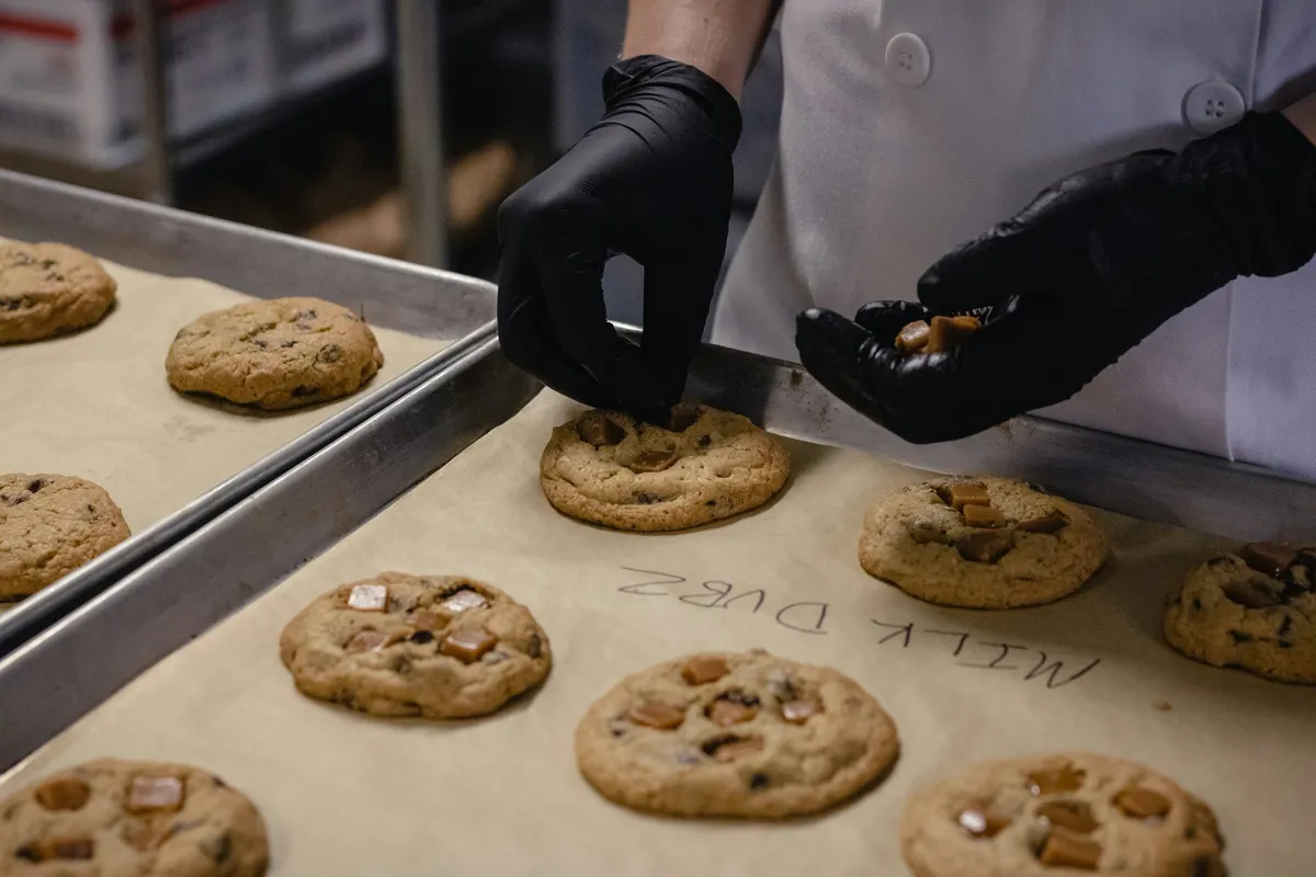 Black-gloved hands press caramel pieces onto chocolate chip cookies on a parchment-lined sheet pan, with a name handwritten on the parchment beside them