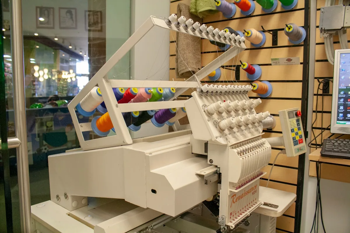 A single-head multi-needle embroidery machine in a shop, with rows of colorful thread cones feeding down into the needle assembly