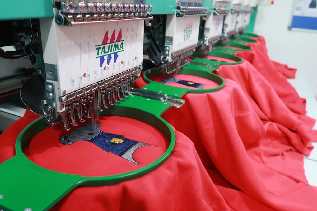 A multi-head commercial embroidery machine stitching a cartoon design into a row of red garments held in green hoops
