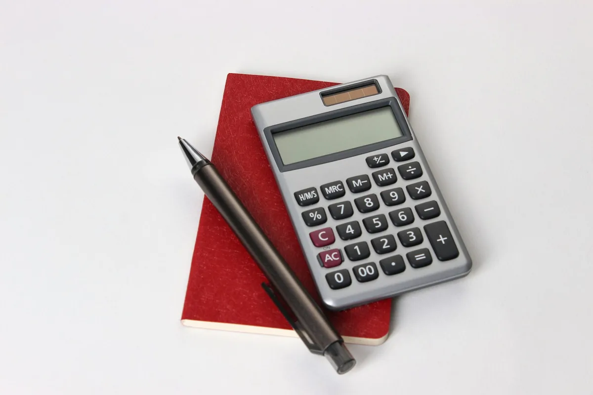 A small calculator and a black pen resting on top of a closed red notebook on a white desk — the visual shorthand for tracking the numbers in a maker business