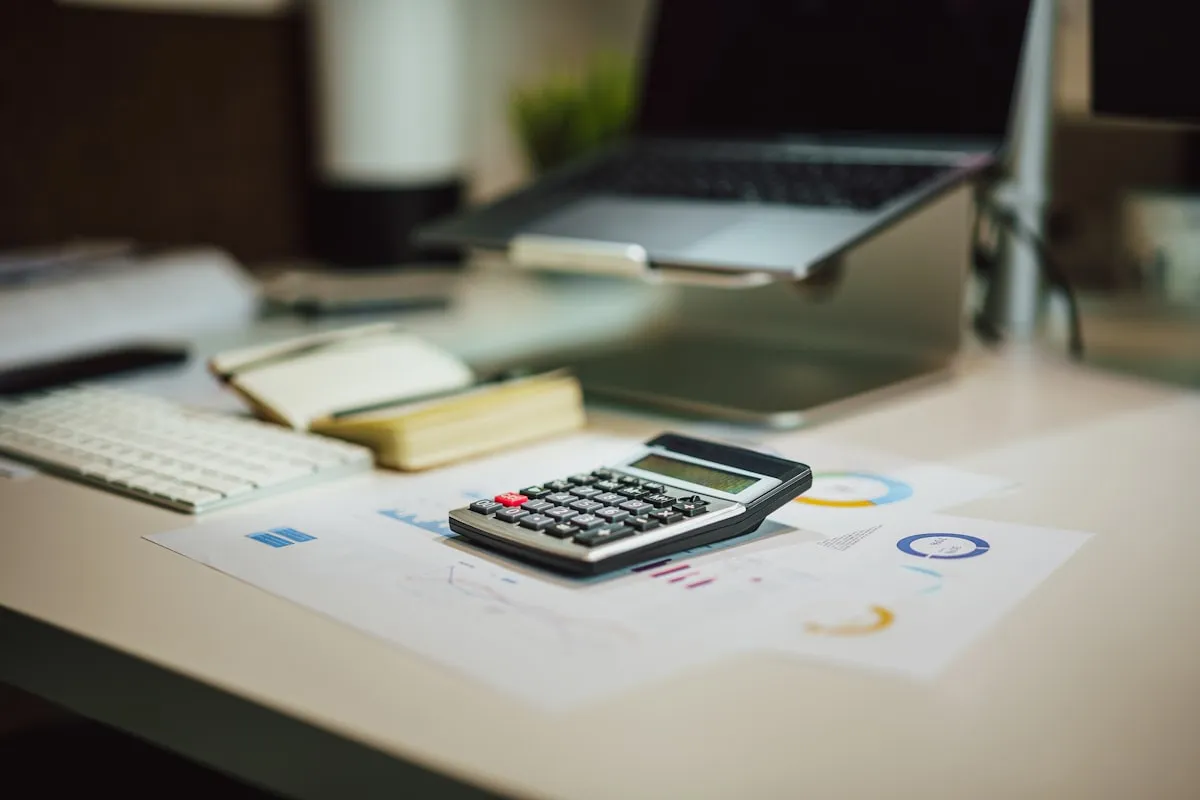 A black calculator resting on printed financial charts on a light wooden desk, with a laptop on a stand and a keyboard blurred in the background