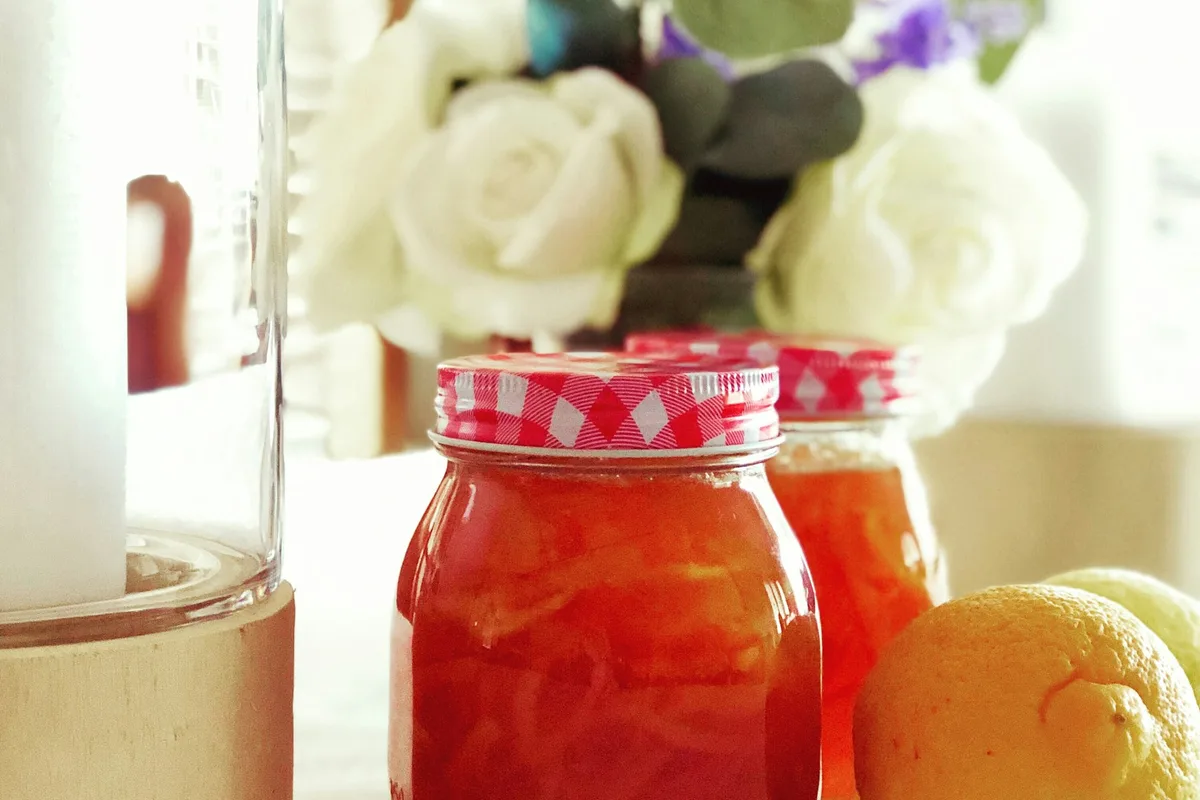 Glass jars of homemade citrus marmalade with red-gingham lids on a sunlit kitchen table beside a fresh lemon and white roses