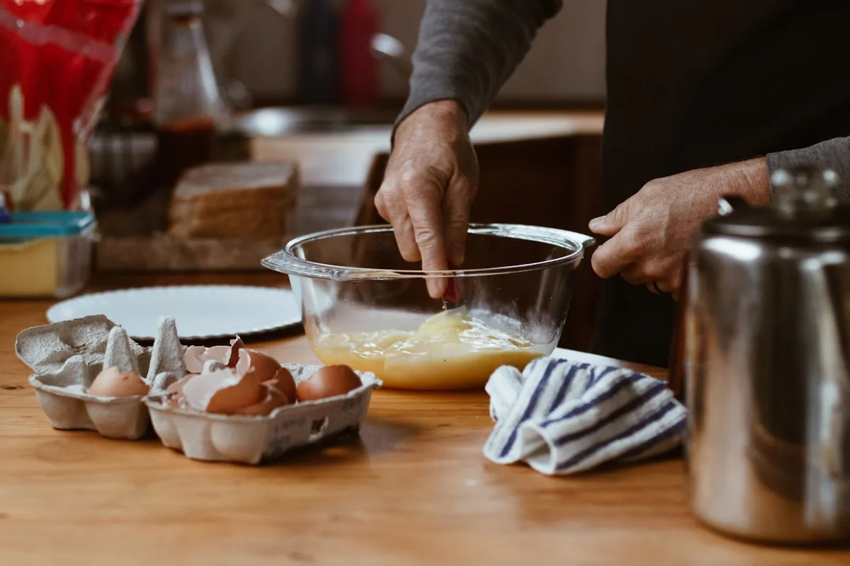 A home baker's hands whisking eggs in a glass mixing bowl on a warm wooden kitchen counter, with an open carton of cracked brown eggs, a folded blue-and-white striped tea towel, and a metal canister nearby, photographed in soft natural light
