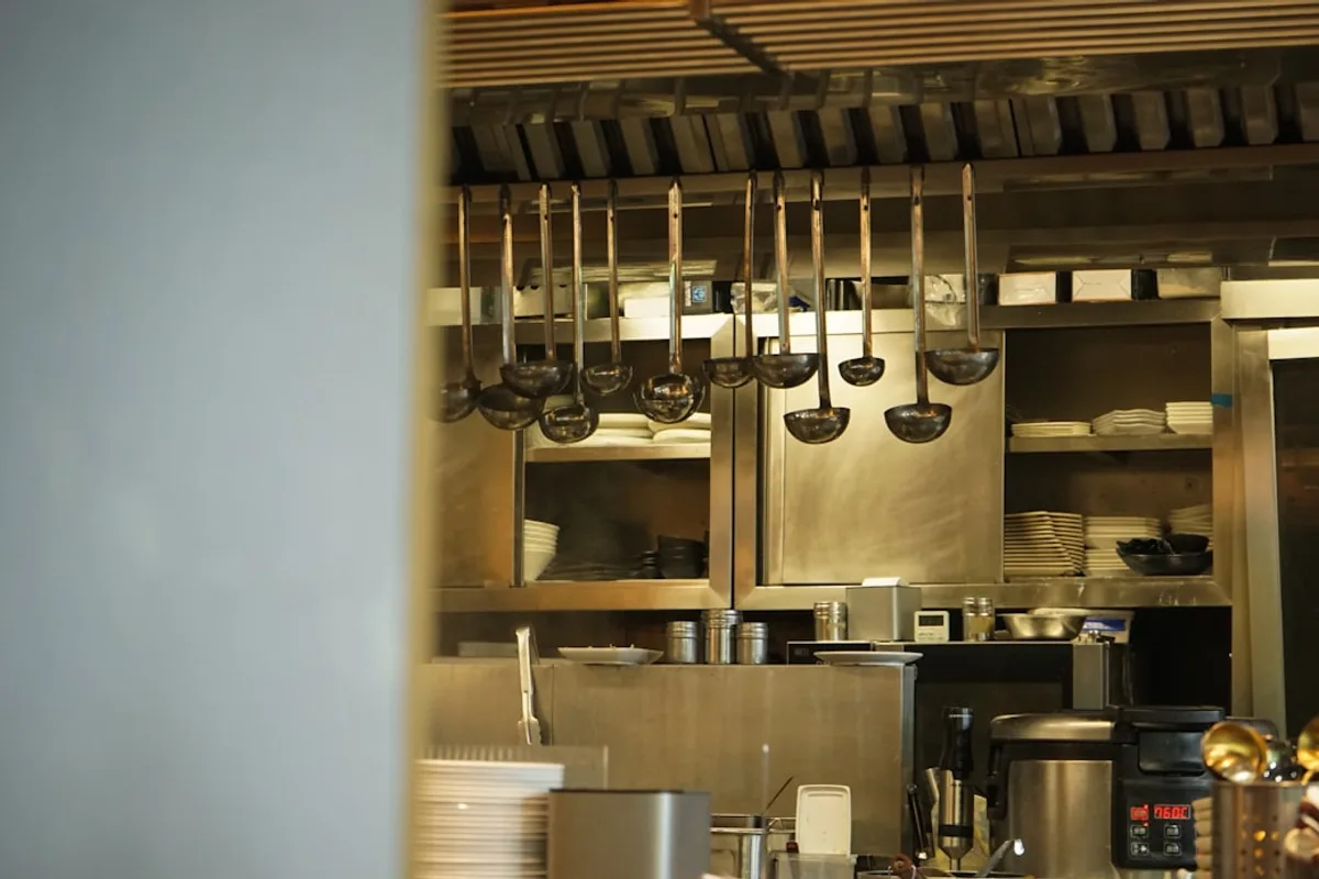 A commercial kitchen line with hanging ladles, stainless steel shelving stacked with plates, and food-service equipment, with no one at the station