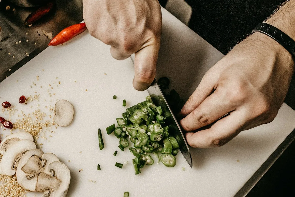 Close-up of two hands chopping green vegetables with a chef's knife on a white cutting board, with sliced mushrooms, diced chili, and raw chicken arranged nearby, prepping ingredients for a batch of meals