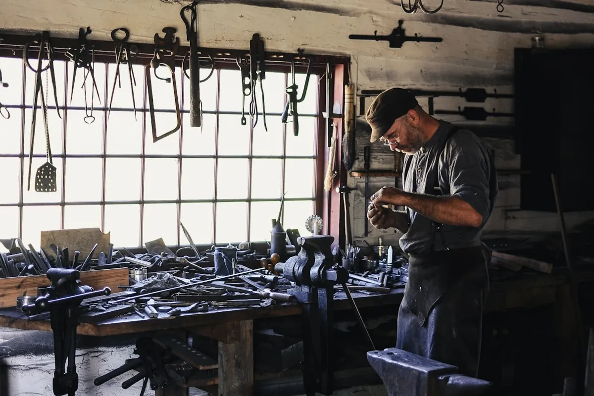A metalworker in a flat cap and leather apron examining a piece at a tool-covered workbench, hand-forged tools hanging by a bright window and an anvil in the foreground