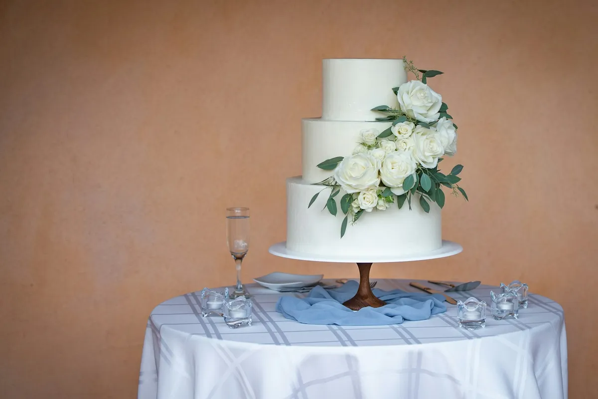 A three-tier white wedding cake decorated with a cascade of fresh white roses and eucalyptus leaves on a wooden cake stand, set on a draped tablecloth with a champagne flute and small glass tea-light holders against a warm terracotta wall