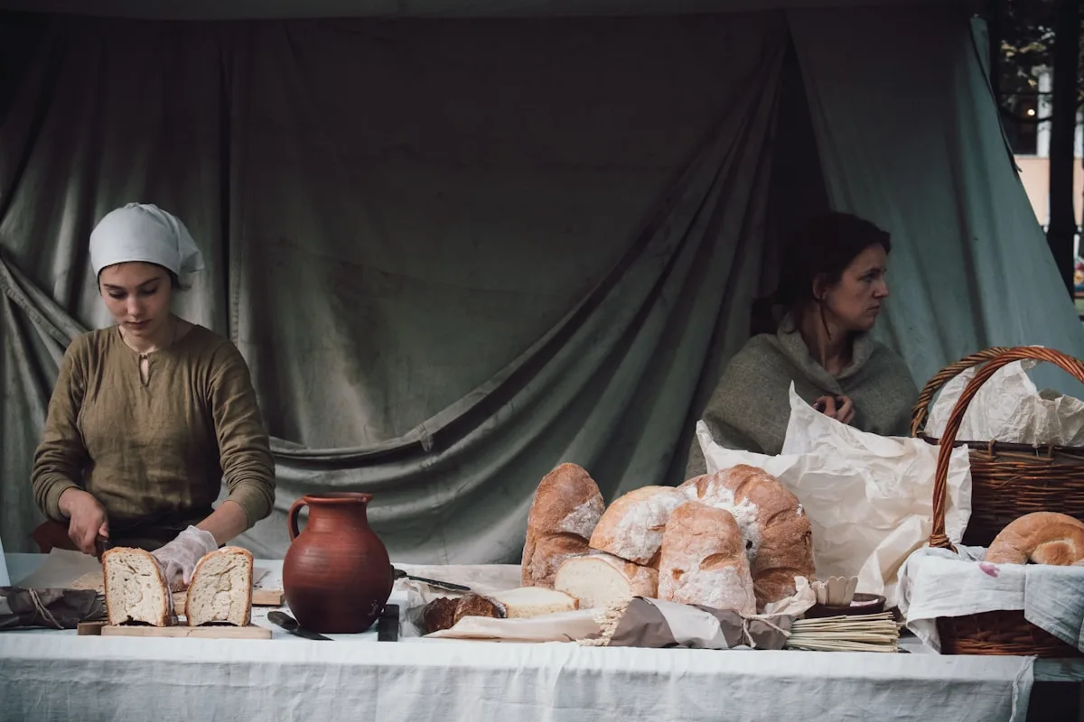 Two bakers selling fresh-baked sourdough loaves at a rustic outdoor market stall — one slicing a loaf with gloved hands beside a clay pitcher, the other tending a basket of breads wrapped in linen, set against a softly draped canvas backdrop in muted daylight